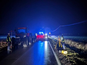 Z daleka ujęcie miejsca po wypadku drogowym. Ciemno, droga, ciągnik rolniczy oraz policjanci ruchu drogowego wykonujący czynności.