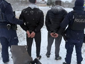 Na zdjęciu w polu stoi dwóch zatrzymanych mężczyzn, których pilnuje dwóch umundurowanych policjantów