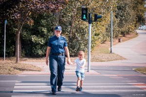 Zdjęcie przedstawiające policjantkę oraz chłopca w trakcie przechodzenia przez przejście dla pieszych.