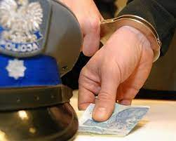 na zdjęciu policyjna czapka oraz ręka skuta w kajdanki. Osoba skuta w kajdanki w dłoni trzyma pieniądze w postaci banknotów.