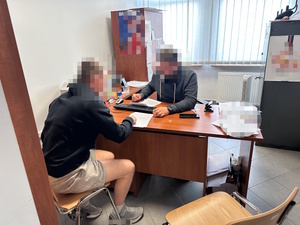 na zdjęciu nieumundurowany policjant wykonuje czynności przesłuchania z zatrzymanym mężczyzną. Oboje siedzą przy biurku.
