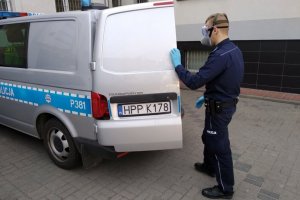 umundurowany policjant ubrany w sprzęt biologicznej ochrony osobistej zamyka drzwi oznakowanego radiowozu - busa