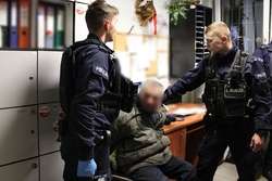 Zdjęcie przedstawia dwóch umundurowanych policjantów w pomieszczeniu policyjnym oraz siedzącego między nimi zatrzymanego mężczyznę.