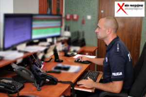 Zdjęcie przedstawia policjanta siedzącego przy biurku i patrzącego w monitory.