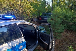 Na zdjęciu widać radiowóz z otwartymi przednimi drzwiami, samochód, którym leśną drogą uciekał nietrzeźwy kierowca, obok stoi umundurowany policjant, który pilnuje zatrzymanego mężczyznę