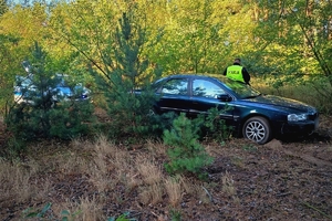 Na zdjęciu widać samochód, którym leśną drogą uciekał nietrzeźwy kierowca, obok stoi umundurowany policjant, w tle radiowóz