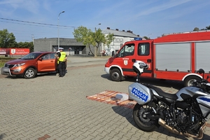 Zdjęcie przedstawia policjanta podejmującego kontrole pojazdu oraz policyjny motocykl i wóz strażacki.