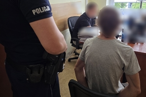 Zdjęcie przedstawia umundurowanego policjanta stojącego obok zatrzymanego siedzącego na krześle oraz nieumundurowanego policjanta przy biurku.