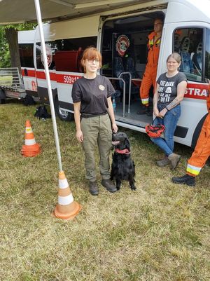 na zdjęciu widać widoczna jest kobieta z Ochotniczej straży pożarnej obok siedzi czarny pies , który jest wykorzystywany do poszukiwania ludzi