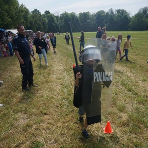 na zdjęciu widoczny jest chłopiec ubrany w kamizelkę policyjna , kask policyjny na głowie w ręce trzyma pałkę policyjna szturmową oraz tarcze z napisem policja , biegnie torem przeszkód