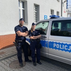 na zdjęciu widoczna jest policjantka i policjant stojący obok oznakowanego radiowozu, za ich plecami widoczny jest budynek komendy .