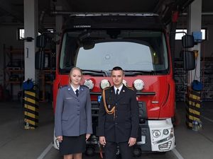 na zdjęciu widoczna jest umundurowana policjantka obok stoi umundurowany strażak z ich plecami widoczny jest wóz strażacki