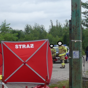 na zdjęciu widoczna jest osłona z napisem straż oraz dwóch umundurowanych strażaków jak również oznakowany radiowóz