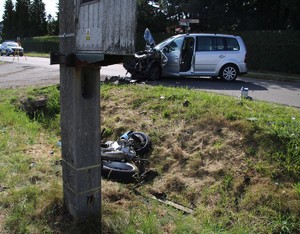 na zdjęciu widoczny jest uszkodzony samochód obok niego na łące leży uszkodzony motocykl widoczne jest również skrzyżowanie ulic