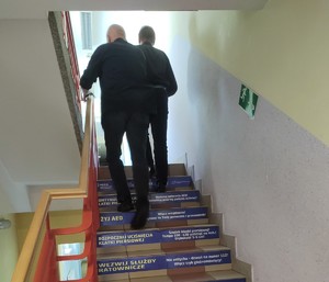 na zdjęciu widocznych jest dwóch mężczyzn ubranych w ciemne kolory idących po schodach w górę na schodach widoczne są naklejki z instrukcja pierwszej pomocy