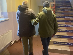 na zdjęciu widocznych jest dwóch mężczyzn jeden z nich ma na nadgarstkach kajdanki drugi trzyma go pod ramie i ma na rękawie opaskę z napisem policja