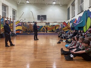 na sali gimnastycznej siedzą na podłodze harcerze przed nimi stoją dwaj policjanci