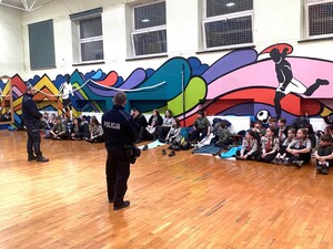 na sali gimnastycznej siedzą na podłodze harcerze przed nimi stoją dwaj policjanci widoczna jest również ściana na której są kolorowe rysunki koszykarza