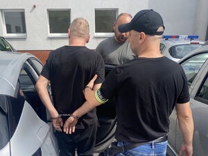mężczyzna stojący tyłem na rękach z tyłu ma kajdanki, obok policjant w ubraniu cywilnym z opaska z napisem policja w tle widoczny drugi policjant ubrany po cywilnemu