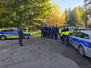 policjanci stoją w dwuszereg obok nich radiowozy przed nimi dwóch umundurowanych funkcjonariuszy za ich plecami widoczny jest las