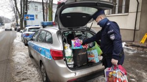 zdjęcie kolorowe: policjant pakuje torby z zabawkami do radiowozu zaparkowanego przed komisariatem