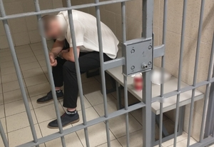 zdjęcie kolorowe: młody mężczyzna ubrany w czarne spodnie i w białą koszule z krótkim rękawem siedzi na ławce w celi aresztu sądowego