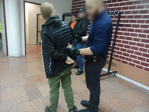 zdjęcie kolorowe: na pierwszym planie nieumundurowany policjant trzymający za rękę zatrzymanego mężczyznę, któremu złożono kajdanki na ręce trzymane z tyłu