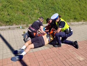 zdjęcie kolorowe: policjantka ruchu drogowego i ratownicy medyczni udzielający pomocy biegaczowi, który zasłabł
