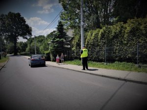 ulica, jedzie samochód osobowy, chodnikiem idą dzieci oraz policjant w odblaskowej kamizelce