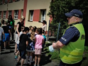 umundurowany policjant stoi na chodniku przed budynkiem szkoły, na drugim planie dzieci oraz budynek placówki