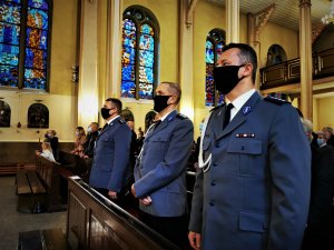 trzech policjantów stoi w ławce kościoła podczas mszy świętej. Usta i nos mają zasłonięte maseczką