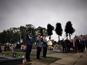 jeden widocznych na zdjęciu policjantów wykonuje gest oddania honoru, policjanci policyjnej orkiestry grają na instrumentach, w tle cmentarz oraz cywilni uczestnicy uroczystości