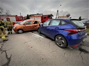 rozbite podczas zdarzenia drogowego samochody, niebieski i pomarańczowy, z tyłu dwa wozy straży pożarnej oraz strażacy