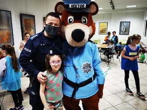 policjant i policyjna maskotka pozują do zdjęcia z dzieckiem. W tle inne dzieci