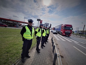 policjanci ruchu drogowego, w odlbaskowych kamizelkach, stoją na chodniku przy trasie drogi krajowej. Widać jadące ulicą samochody