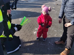 na zdjęciu widać małą dziewczynkę w różowym kombinezonie zimowym, policjantka wręcza jej mamie odblaskowe opaski