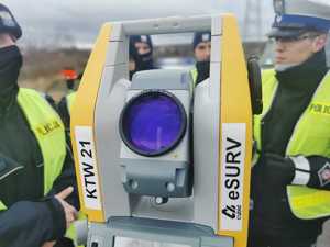 zdjęcie przedstawia urządzenie zwane tachimetrem, w tle policjanci drogówki