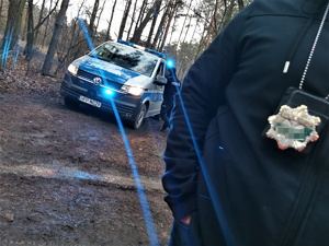 na zdjęciu widać policyjny radiowóz, w tle las, na pierwszym planie fragment kurtki policjanta, który na szyji na przewieszoną policyjną odznakę
