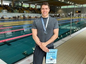 zdjęcie przedstawia mężczyznę z medalem na szyi trzymającego dyplom, mężczyzna stoi na basenie.