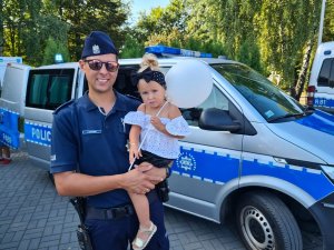 Umundurowany policjant trzymający dziecko na rękach na tle radiowozu