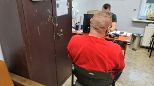 Zatrzymany mężczyzna siedzący na krześle podczas przesłuchania. Przy biurku siedzi nieumundurowany policjant
