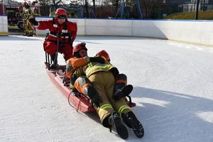 Pokaz ratowania osoby na lodzie w wykonaniu Straży Pożarnej. Osoby znajdują się na saniach ratowniczych na płycie lodowiska.