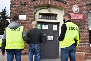 Dwaj umundurowani policjanci w kamizelkach odblaskowych z napisem &quot;Policja&quot; stoją przed budynkiem prokuratury z mężczyzną, który ma założone kajdanki zespolone .