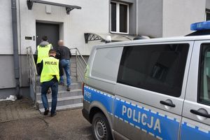 Dwaj umundurowani policjanci w kamizelkach odblaskowych z napisem &quot;Policja&quot; wchodzą do budynku Komendy Miejskiej Policji w Piekarach Śląskich z mężczyzną, który ma założone kajdanki zespolone . Z przodu stoi oznakowany radiowóz