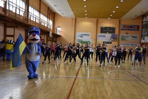 Uczestnicy zajęć sportowo-tanecznych ćwiczący na hali sportowej. Z boku stoi maskotka Śląskiej Policji