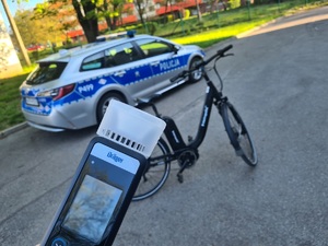 Zdjęcie przedstawia alkomat na tle roweru oraz oznakowanego radiowozu