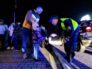 Zdjęcie kolorowe: umundurowany policjant daje dziewczynce opaskę odblaskową, obok niej stoi mężczyzna. W tle widoczni inni uczestnicy