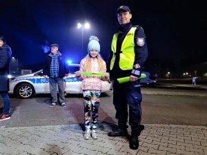 Zdjęcie kolorowe: umundurowany policjant stoi obok dziewczynki, która w rękach trzyma opaskę odblaskową, w tle widoczny radiowóz