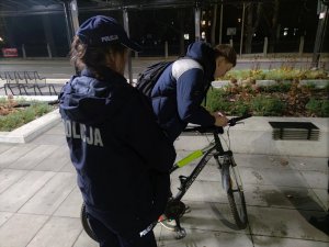 Zdjęcie kolorowe: na pierwszym planie umundurowana policjantka. Następnie widoczny młody mężczyzna, który zapina lampkę rowerową.