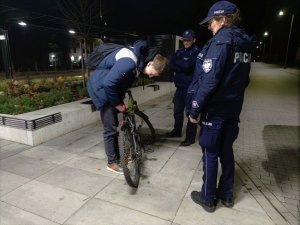Zdjęcie kolorowe: umundurowani policjanci stoją obok młodego mężczyzny, który zapina do roweru lampkę rowerową z tyłu